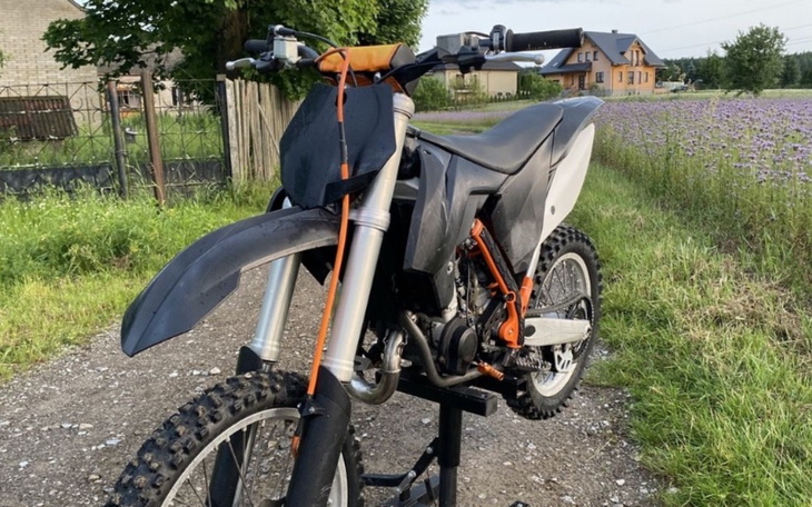 Motocykl terenowy na stojaku, w otoczeniu zieleni i domów.