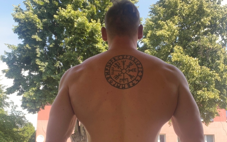 Mężczyzna z tatuażem na plecach, stojący na tle drzew.