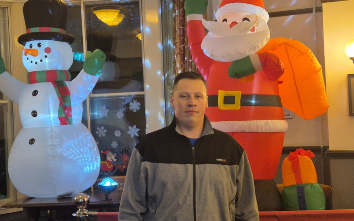 Mężczyzna w szarej bluzie, stoi obok dekoracji świątecznych.