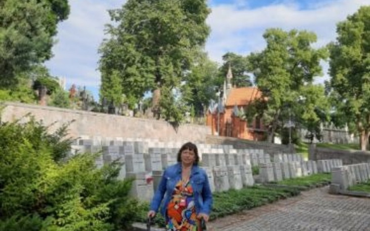 Kobieta w kolorowej sukience na tle cmentarza z drzewami.