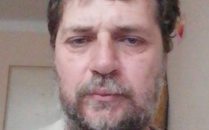 Mężczyzna z zarostem, patrzący w obiektyw, w tle jasne pomieszczenie.