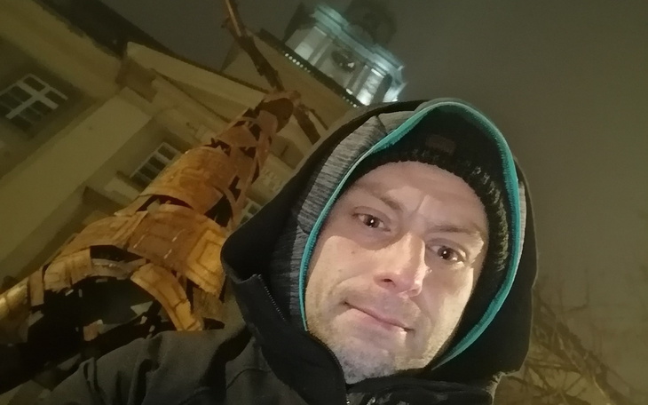 Mężczyzna w kapturze, w tle widoczny budynek w nocy.