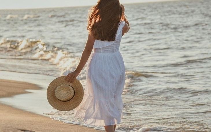 Kobieta w białej sukience spacerująca po plaży, trzymająca kapelusz.