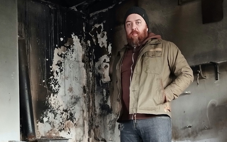Mężczyzna w zniszczonym pomieszczeniu, z brodą i czapką, w stylu casual.