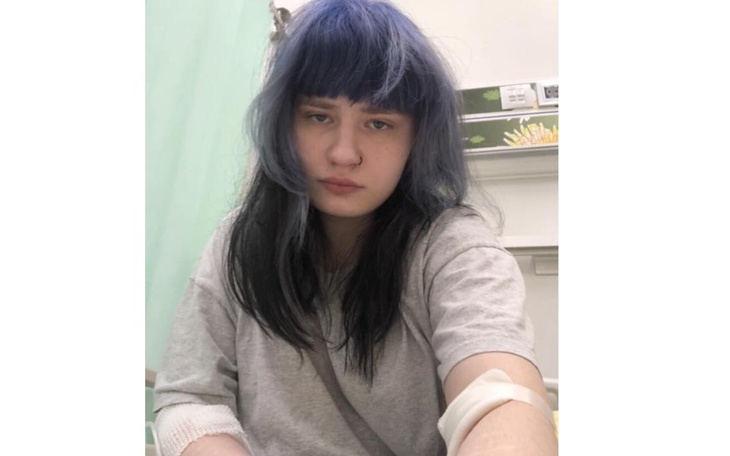 Młoda osoba w szpitalnej koszuli, z niebieskimi włosami, wyglądająca na zmęczoną.