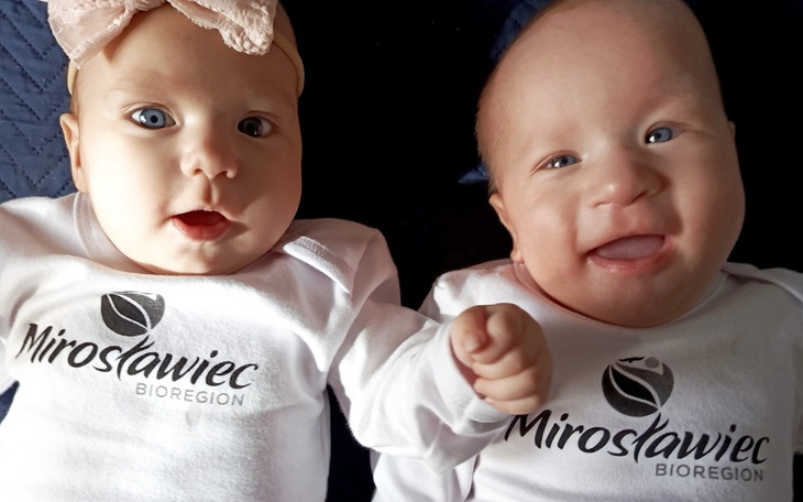 Dwoje niemowląt w białych body, uśmiechających się do kamery.