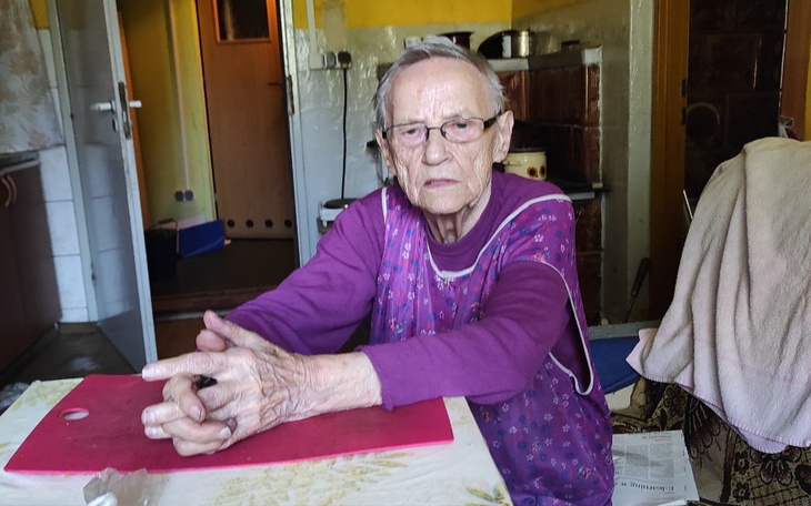 Starsza kobieta siedzi przy stole, z rękami na blacie, w kuchni.