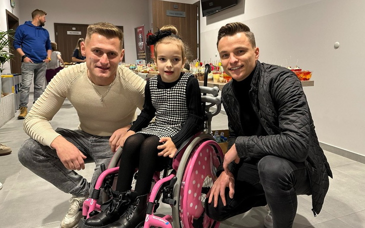 Trzech ludzi, w tym dziewczynka na wózku, uśmiechających się do kamery.