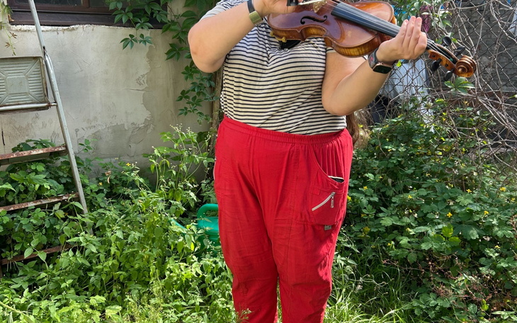Osoba grająca na skrzypcach w ogrodzie, ubrana w paski i czerwone spodnie.