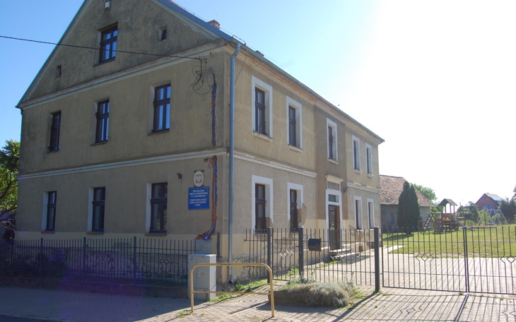 Zabytkowy budynek z ogrodzeniem i zielenią w tle.