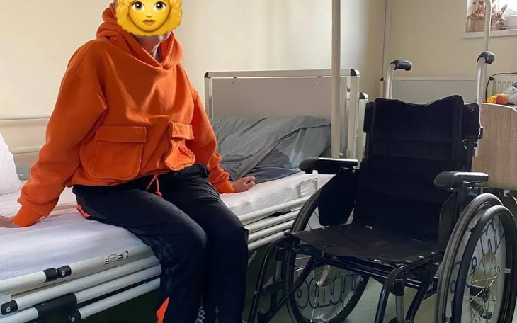 Osoba w pomarańczowej bluzie siedząca na łóżku w szpitalu obok wózka inwalidzkiego.