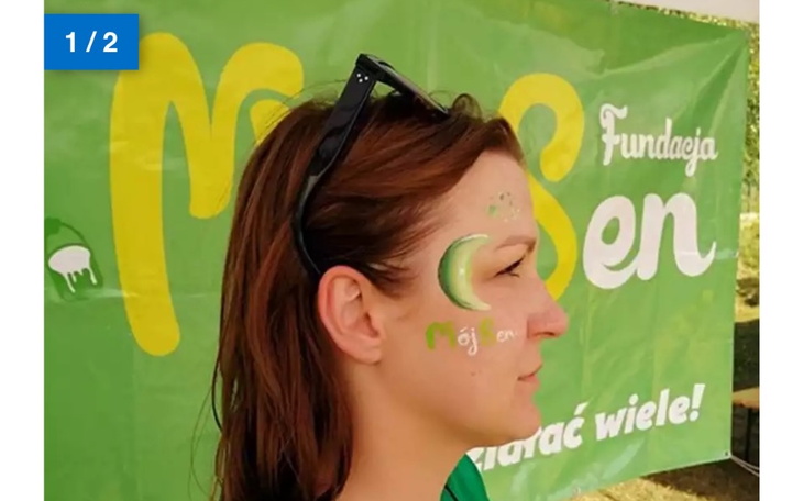 Kobieta z malowidłem na twarzy, stojąca przed zielonym tłem z napisem.