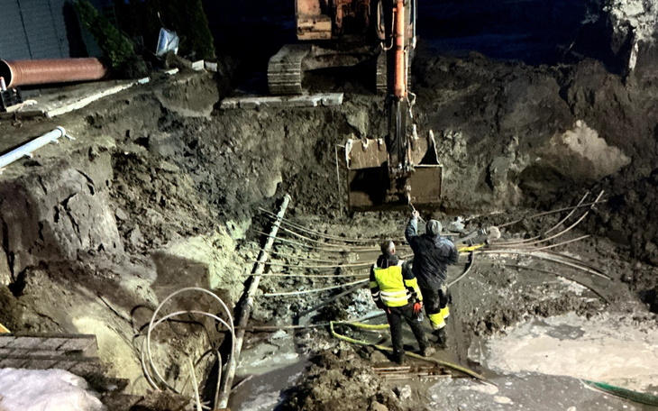 Ekipa budowlana pracująca w wykopie, z maszyną w tle.