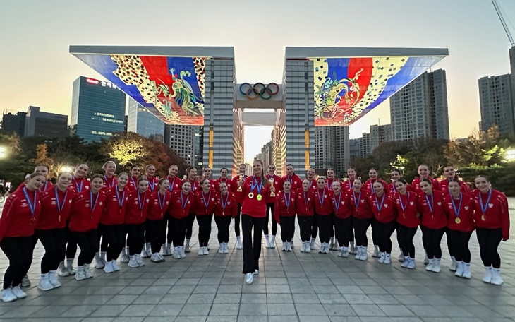 Grupa sportowców w czerwonych kurtkach z medalami na tle olimpijskim.