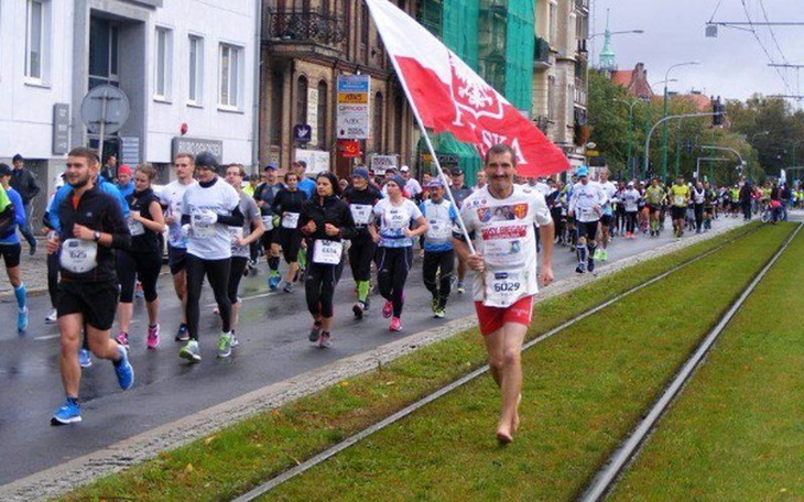Biegacze na ulicy, w tym jeden z flagą, podczas wydarzenia biegowego.