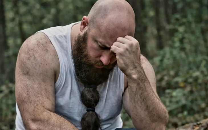 Mężczyzna z brodą siedzi w lesie, wygląda na zamyślonego.