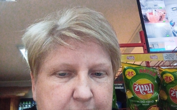 Kobieta w sklepie, z blondu włosami, w tle produkty na półkach.