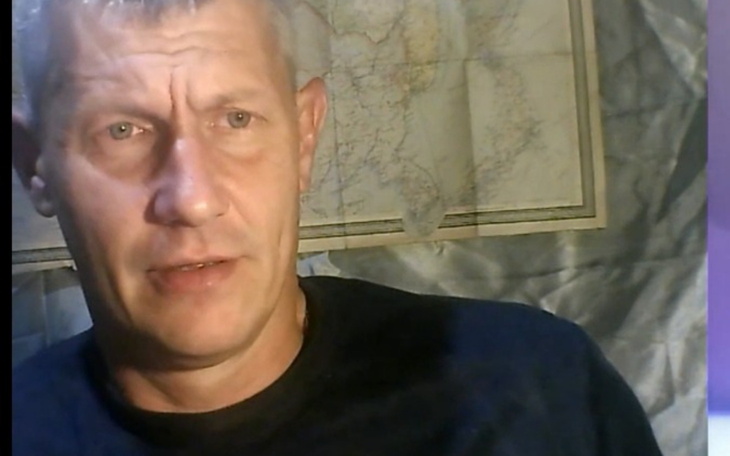 Mężczyzna w koszulce, siedzący przed mapą w tle.