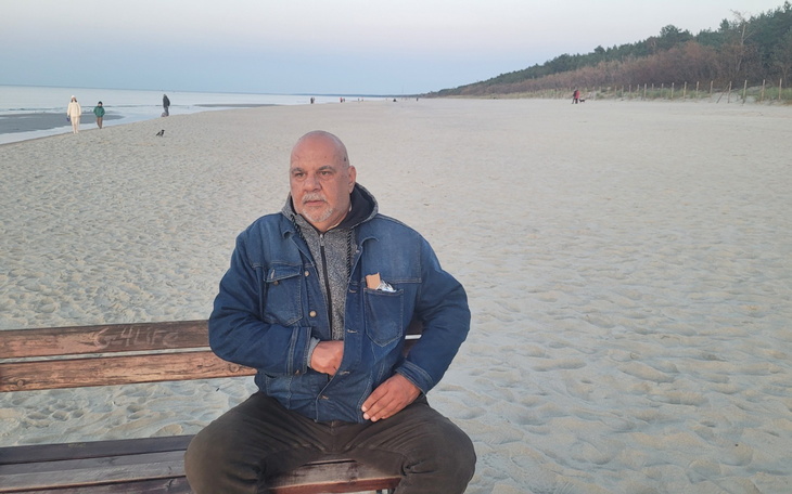 Mężczyzna siedzący na ławce na plaży, w tle morze i las.