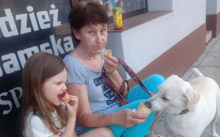 Dziecko i starsza kobieta jedzą lody z psem obok.