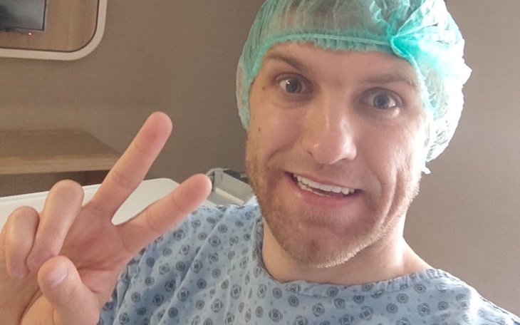 Mężczyzna w szpitalnym fartuchu, uśmiechający się i pokazujący znak pokoju.