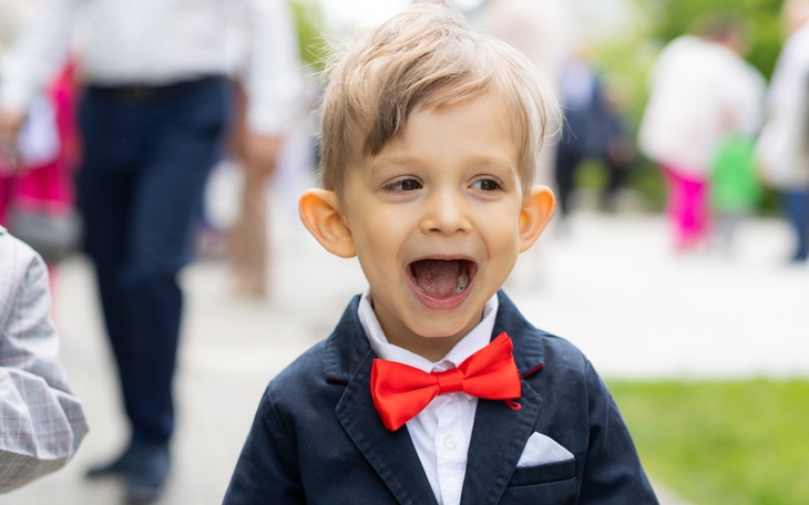 Chłopiec w eleganckim garniturze z czerwoną muszką, uśmiechający się.