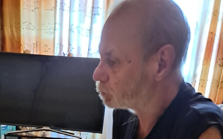 Mężczyzna z brodą, siedzący w pokoju z telewizorem w tle.