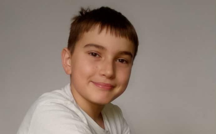 Uśmiechnięty chłopiec z krótkimi włosami, siedzący na tle jasnej ściany.