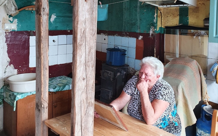 Starsza kobieta siedzi przy stole w kuchni, zamyślona nad czymś.