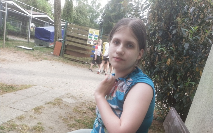 Dziewczyna siedzi na ławce, w tle zieleń i ludzie.