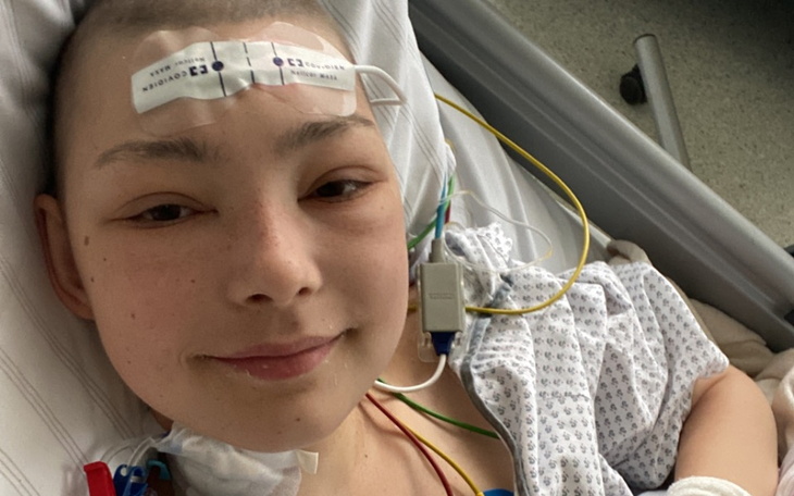 Młoda osoba w szpitalnym łóżku, uśmiechająca się do kamery.