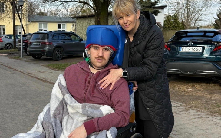 Młody mężczyzna na wózku inwalidzkim, z opiekunką w tle.