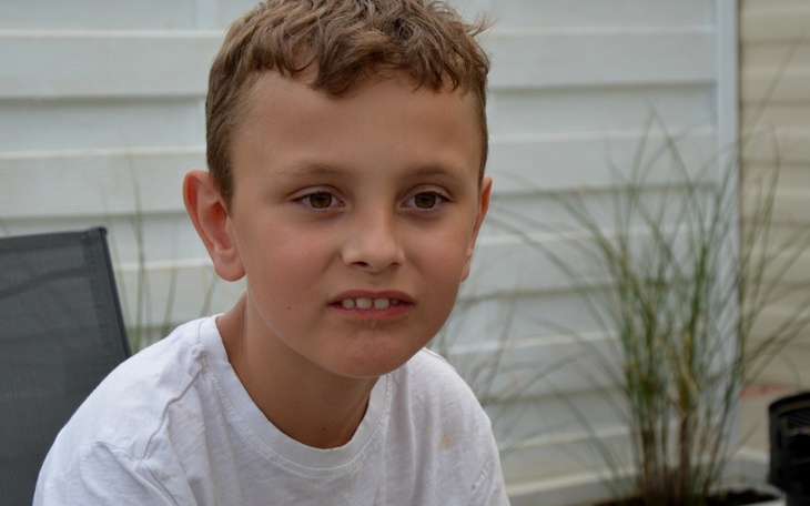 Chłopiec w białej koszulce, z zamyśloną miną, siedzi na zewnątrz.