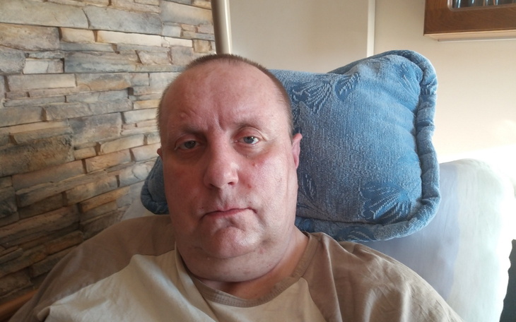 Mężczyzna w szarej koszulce, siedzący na łóżku z poduszką.