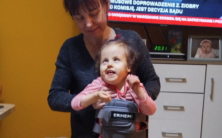 Kobieta wspiera dziecko w specjalnym urządzeniu rehabilitacyjnym.