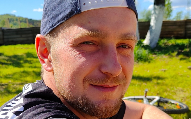 Mężczyzna w kapeluszu, uśmiechający się na tle zieleni.