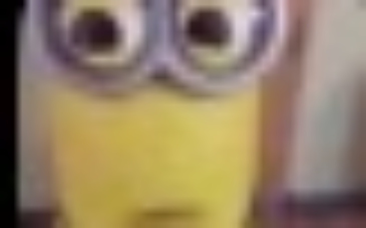 Minionek z dużymi oczami, stojący na tle nieostrego tła.