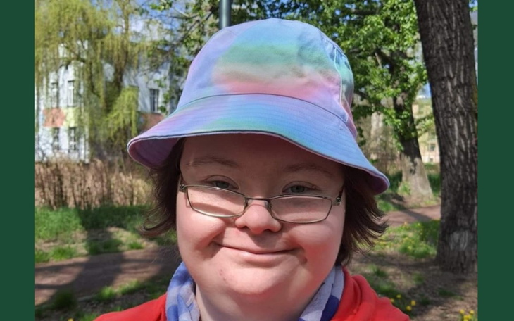 Młoda osoba w kolorowym kapeluszu uśmiecha się na tle zieleni.