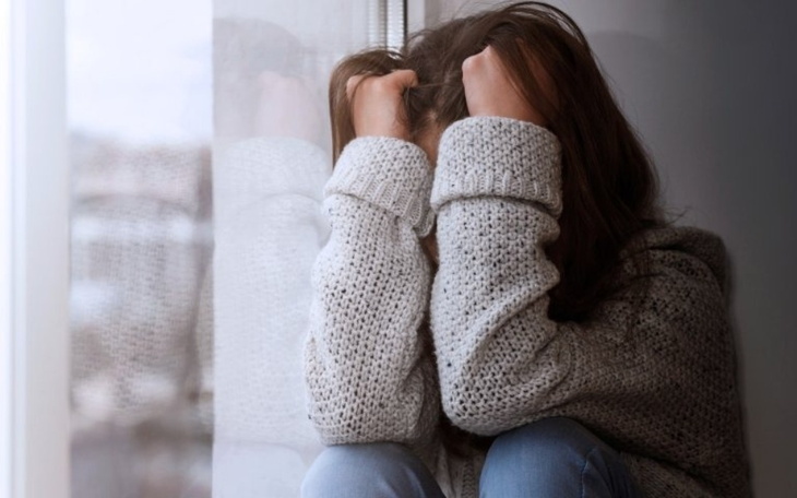 Dziewczyna w szarej bluzie siedzi przy oknie, trzymając głowę w dłoniach.