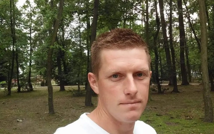 Mężczyzna w lesie, patrzy w obiektyw z poważnym wyrazem twarzy.