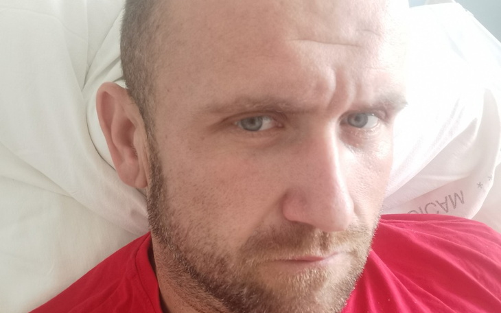 Mężczyzna w czerwonej koszulce, z poważnym wyrazem twarzy, leży na łóżku.