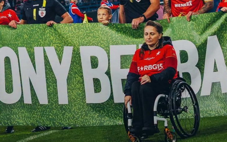 Osoba na wózku z czerwonym logo, trzyma banner i stoi na zielonej murawie.