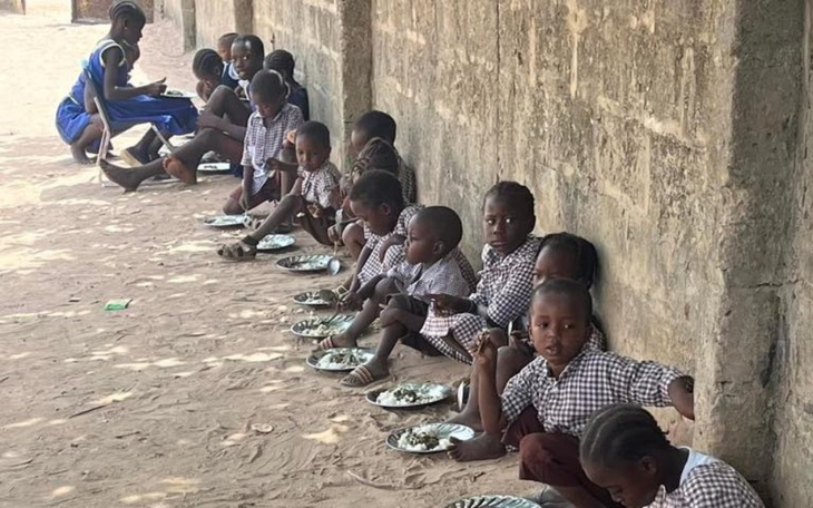 Dzieci jedzące posiłek na zewnątrz, siedzące wzdłuż ściany.