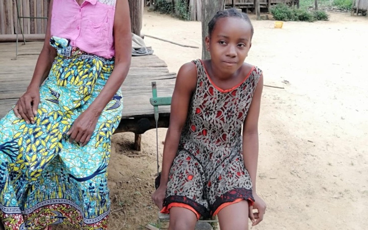 Dziecko w kolorowej sukience siedzi obok dorosłej osoby na drewnianym stołku.