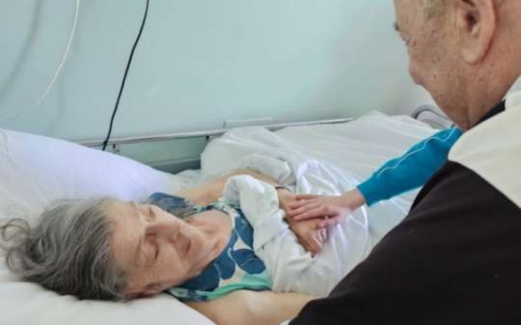 Starsza kobieta w szpitalnym łóżku, trzymana za rękę przez bliską osobę.