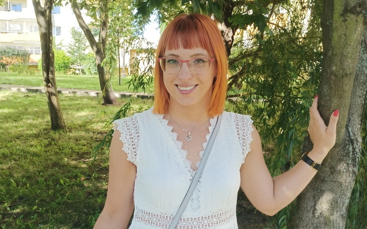 Kobieta w białej sukience stoi przy drzewie, uśmiechając się.