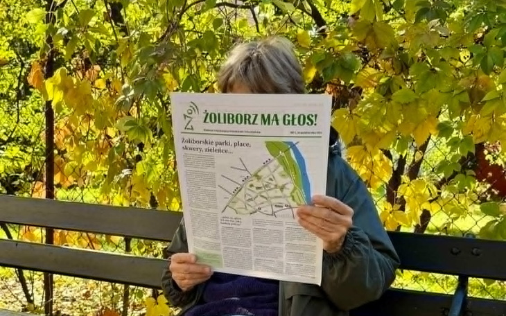 Osoba czytająca gazetę w parku, otoczona kolorowymi liśćmi.