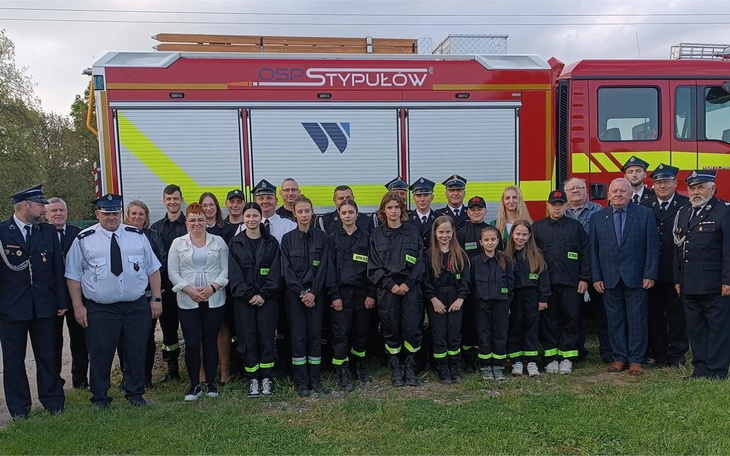 Grupa strażaków w mundurach, pozująca przed wozem strażackim.