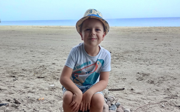 Chłopiec w kapeluszu siedzi na plaży, uśmiechając się do kamery.