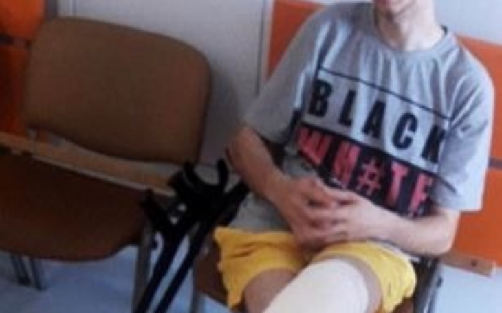 Młody mężczyzna w szarej koszulce siedzi na krześle, z opatrunkiem na nodze.